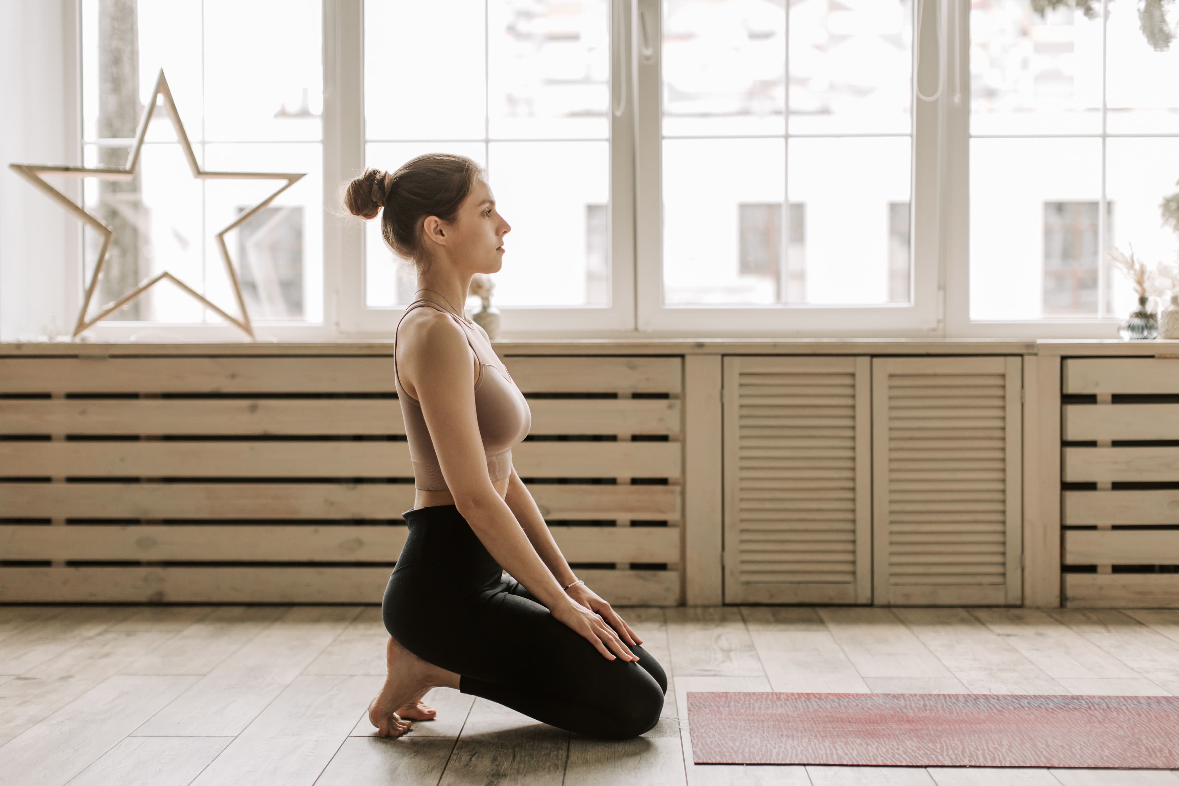 A woman kneeling upright on the floor of a sunlit studio in a calm, still pose.