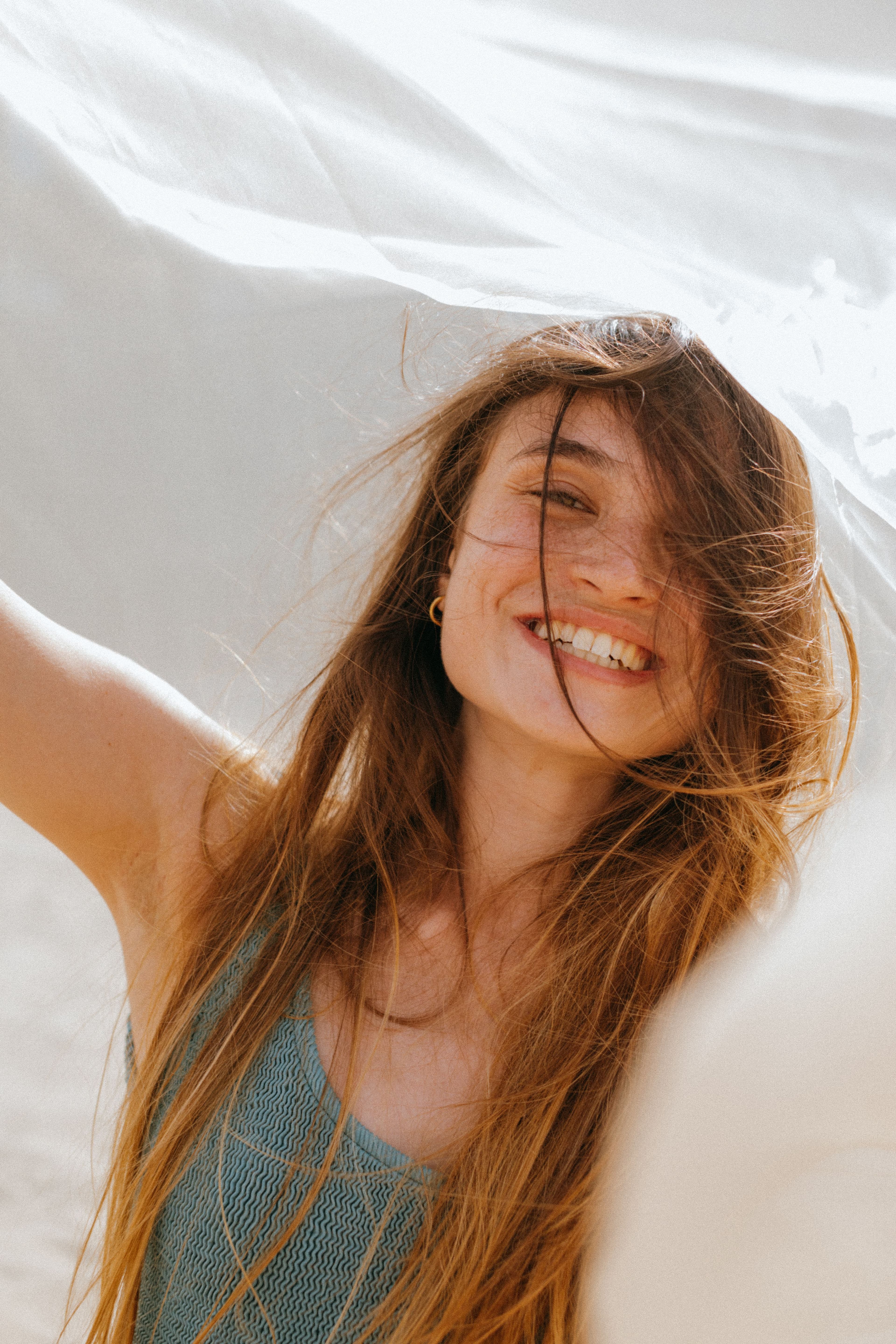A smiling woman with long, windswept hair, one arm raised beneath a billowing sheer white fabric in warm sunlight.