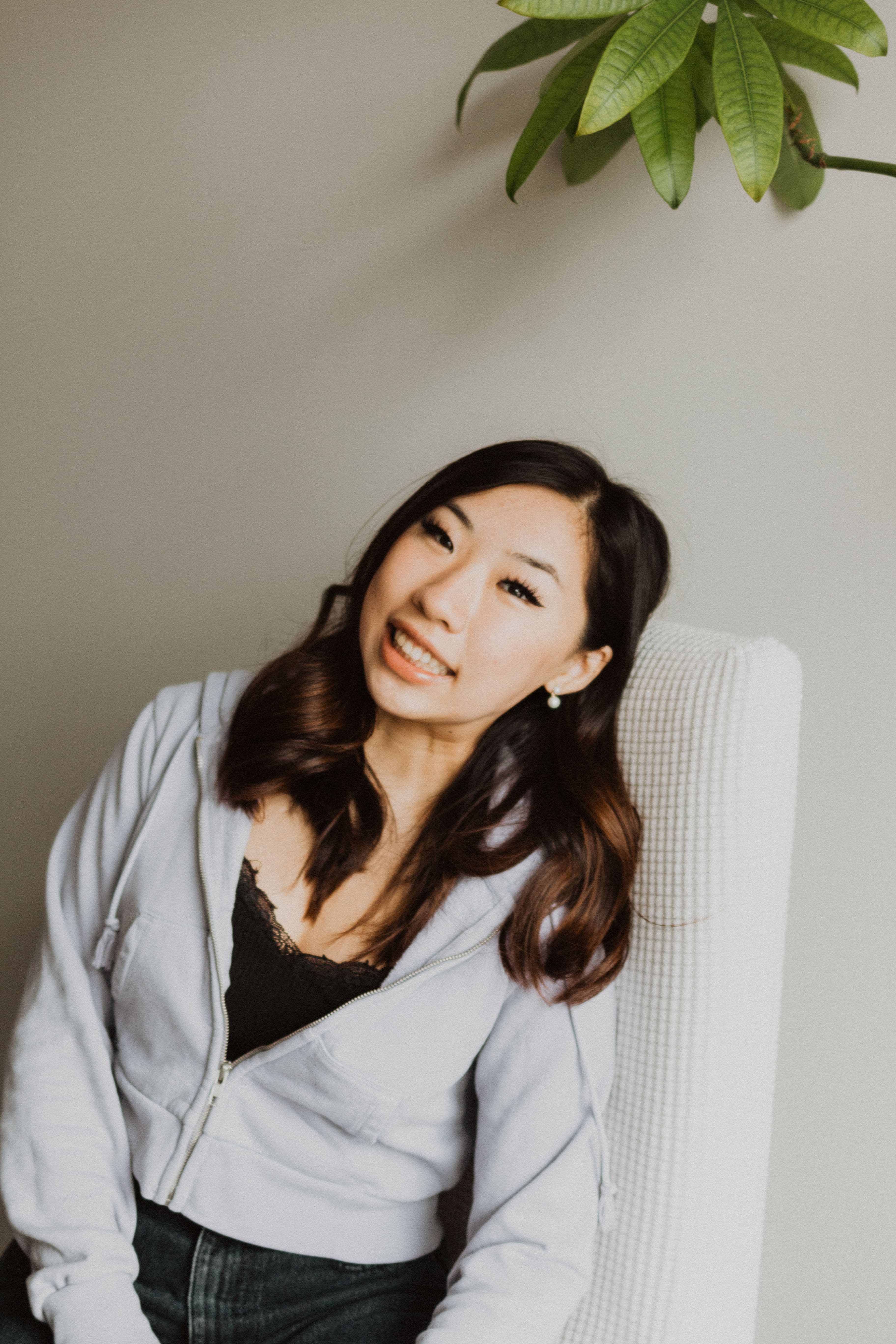 Portrait of a smiling woman with dark hair, seated against a soft grey wall beneath a leafy plant.