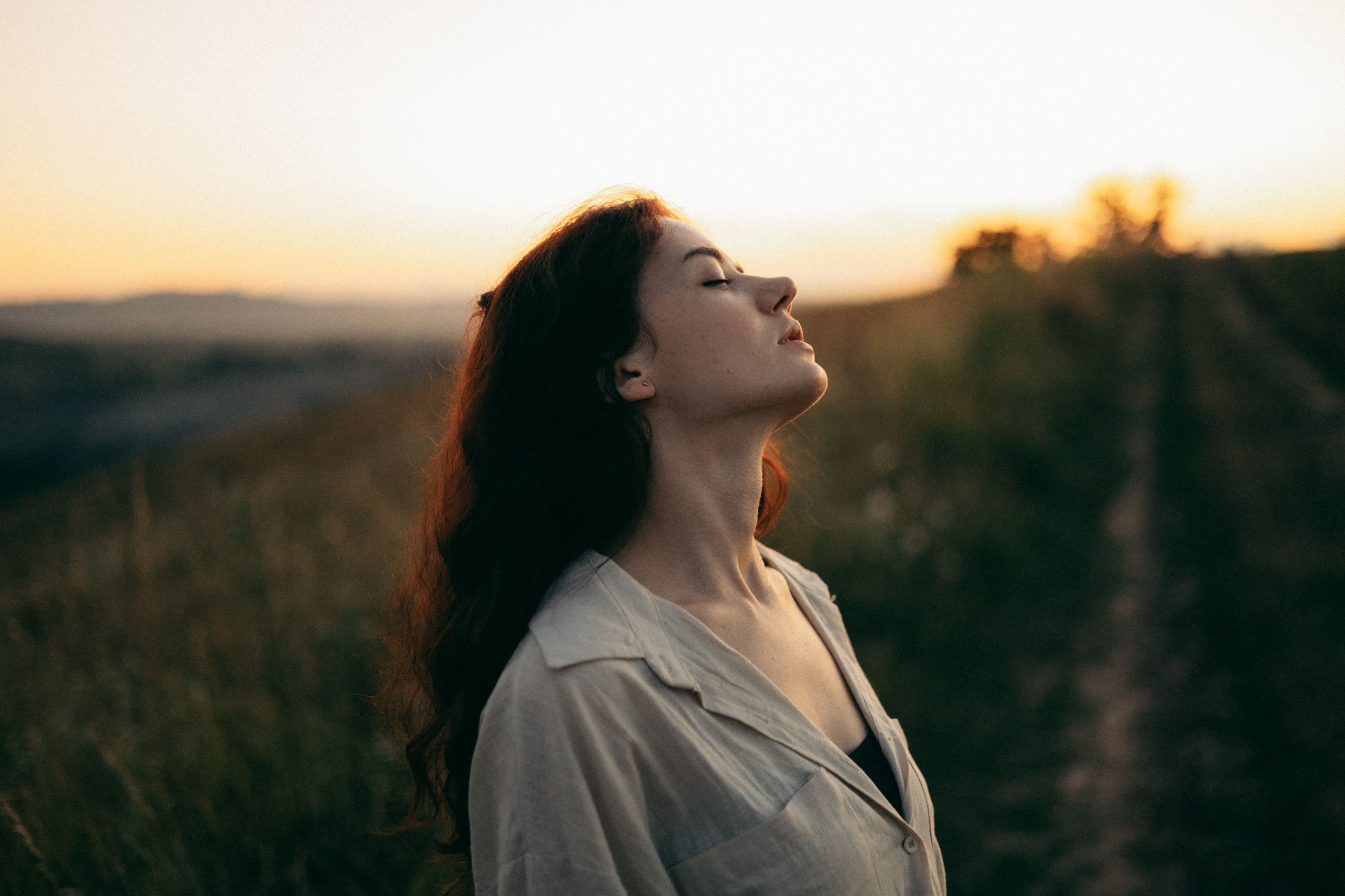 A woman with her head tilted back and eyes closed, breathing in warm evening light in an open field.