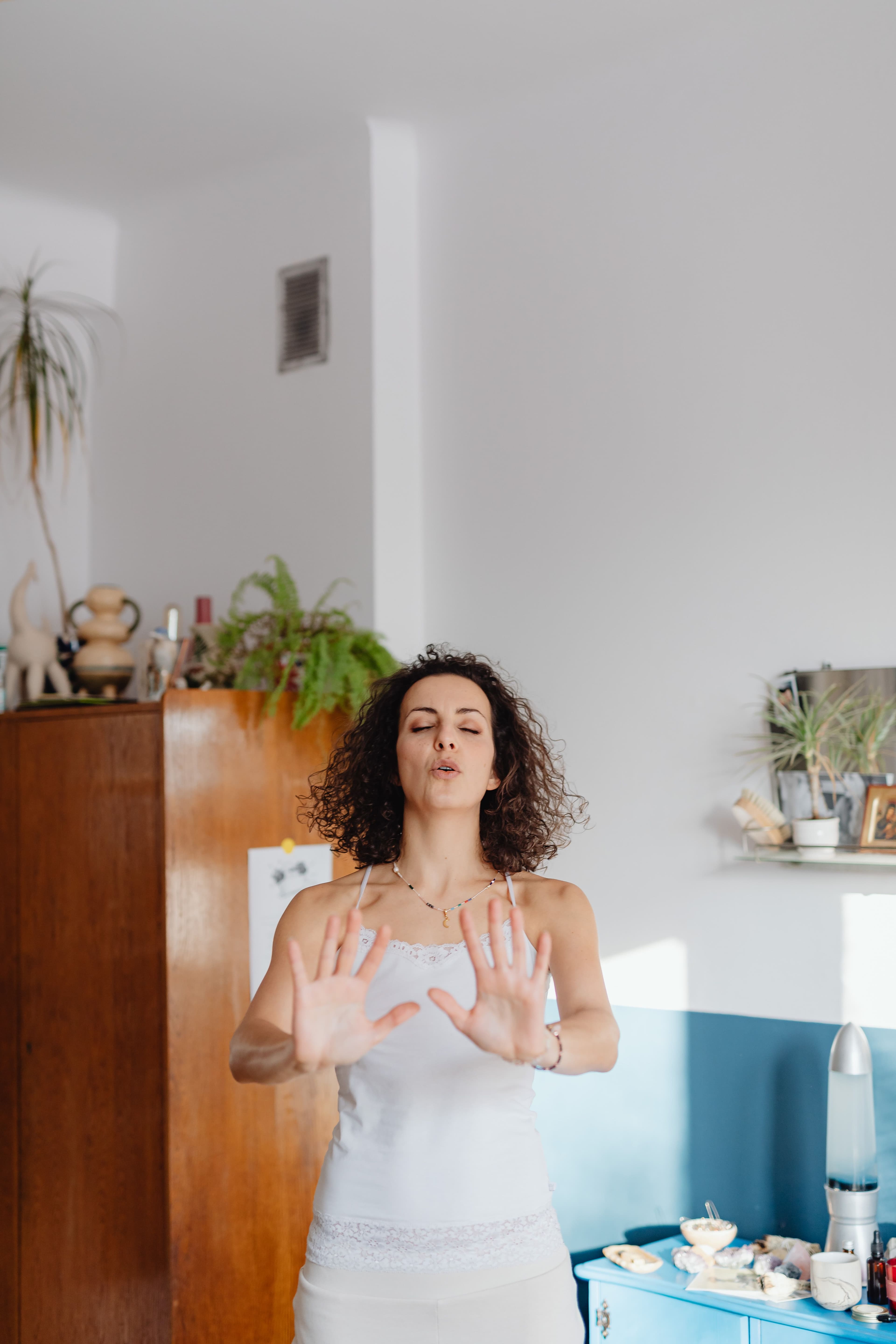 A woman with curly hair standing in a bright living room with her eyes closed and her palms raised, breathing calmly.