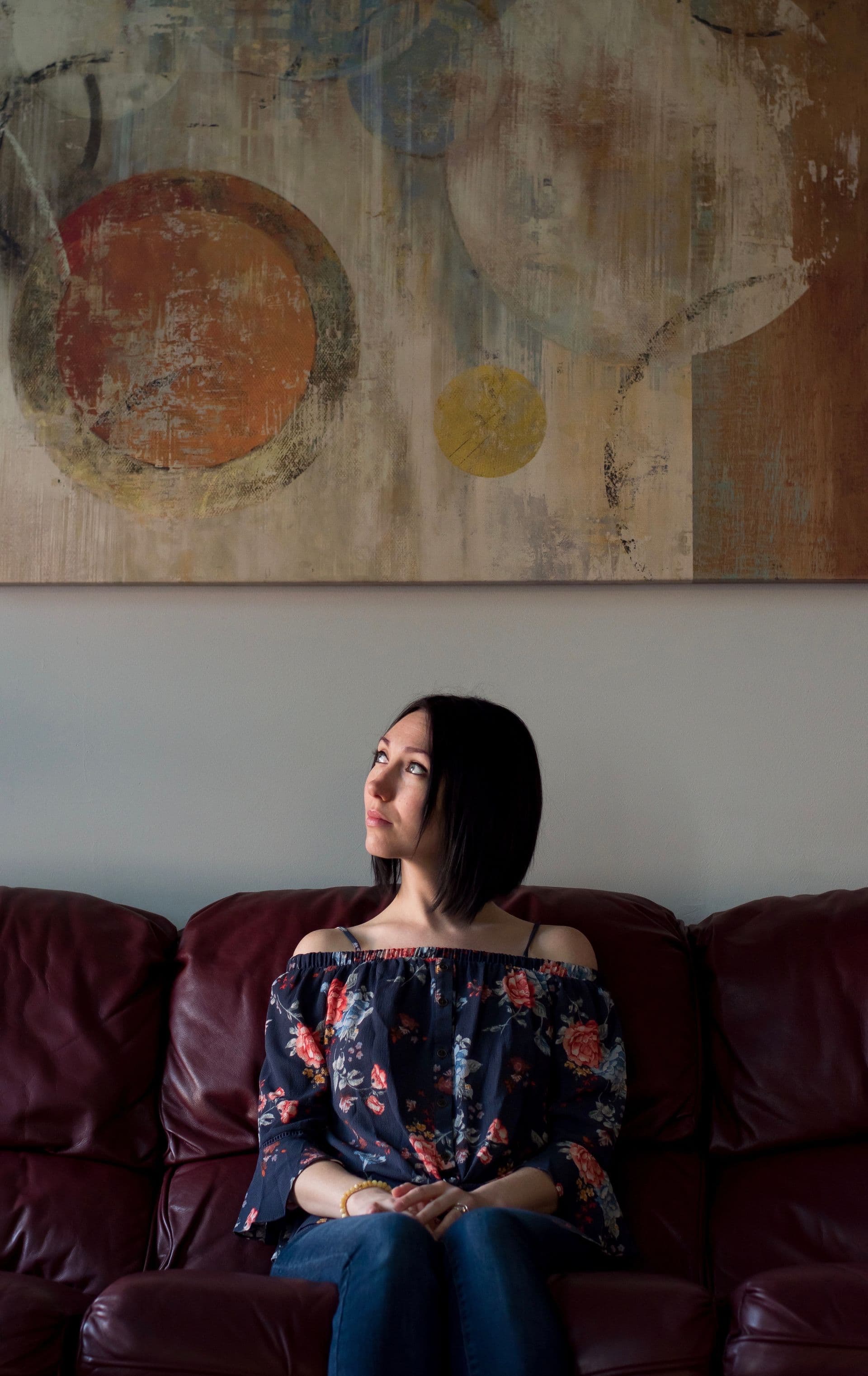A woman with short dark hair sitting on a leather sofa beneath a large abstract painting, looking upward with a thoughtful expression.