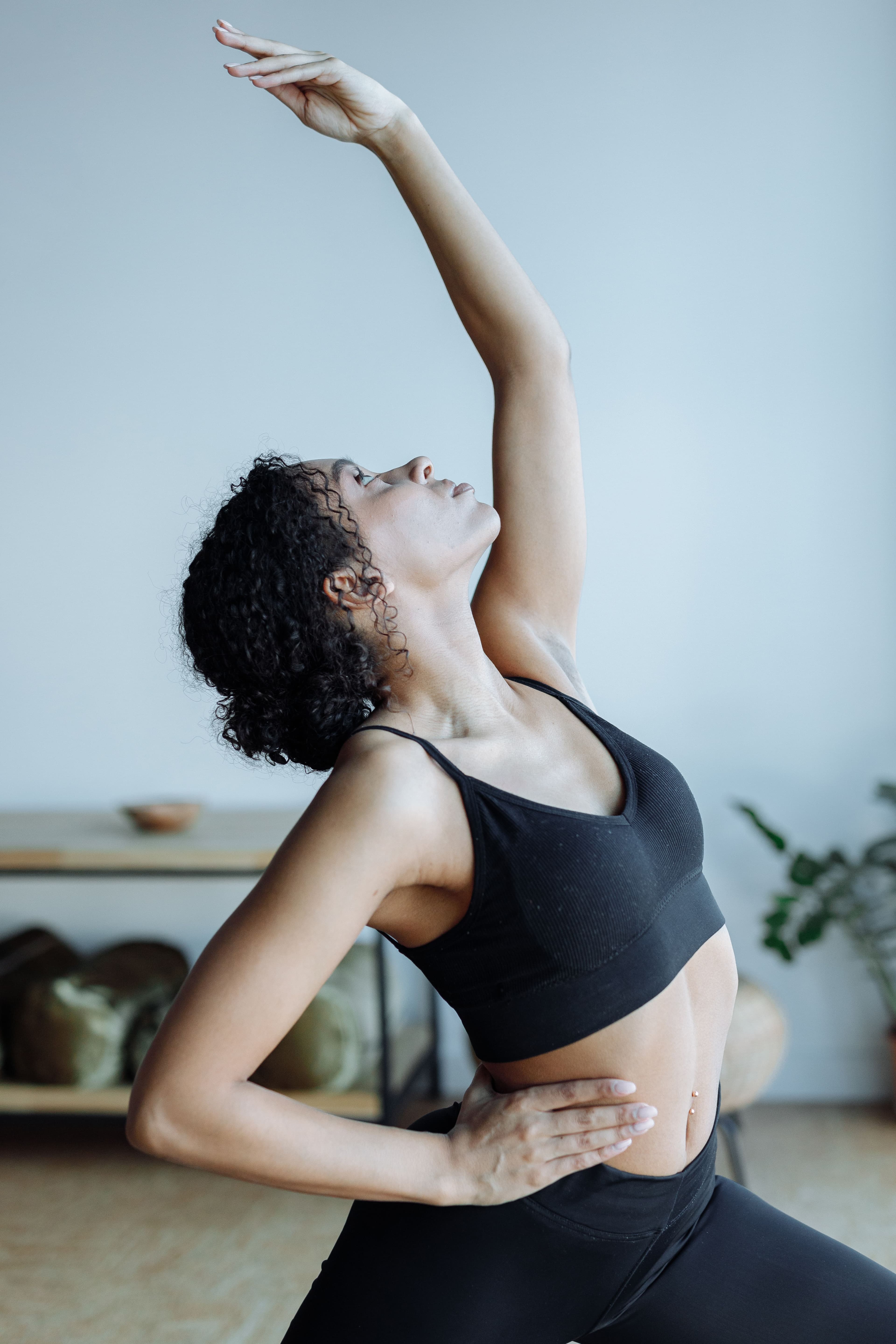 A woman with curly hair in a lunge, one arm reaching high and her head tilted back, in a bright room with a soft blue wall.