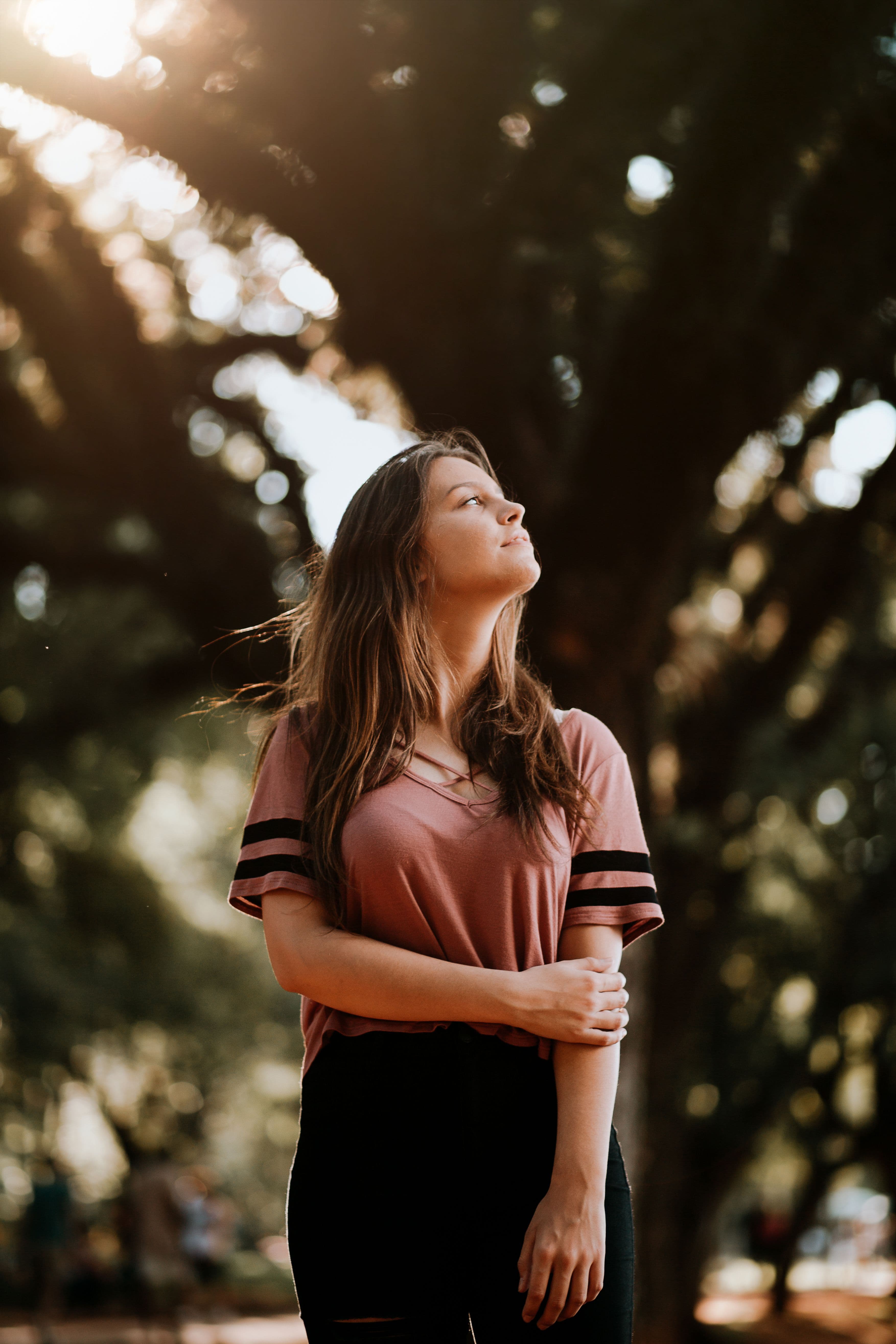 A young woman standing among trees in warm sunlight, looking upward with a calm expression.